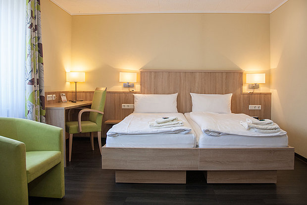 Doppelbett mit weißer Bettwäsche, Lampen rechts und links, und Ablage, Holzbett in der Mitte, links Fenster und ein grüner Sessel
